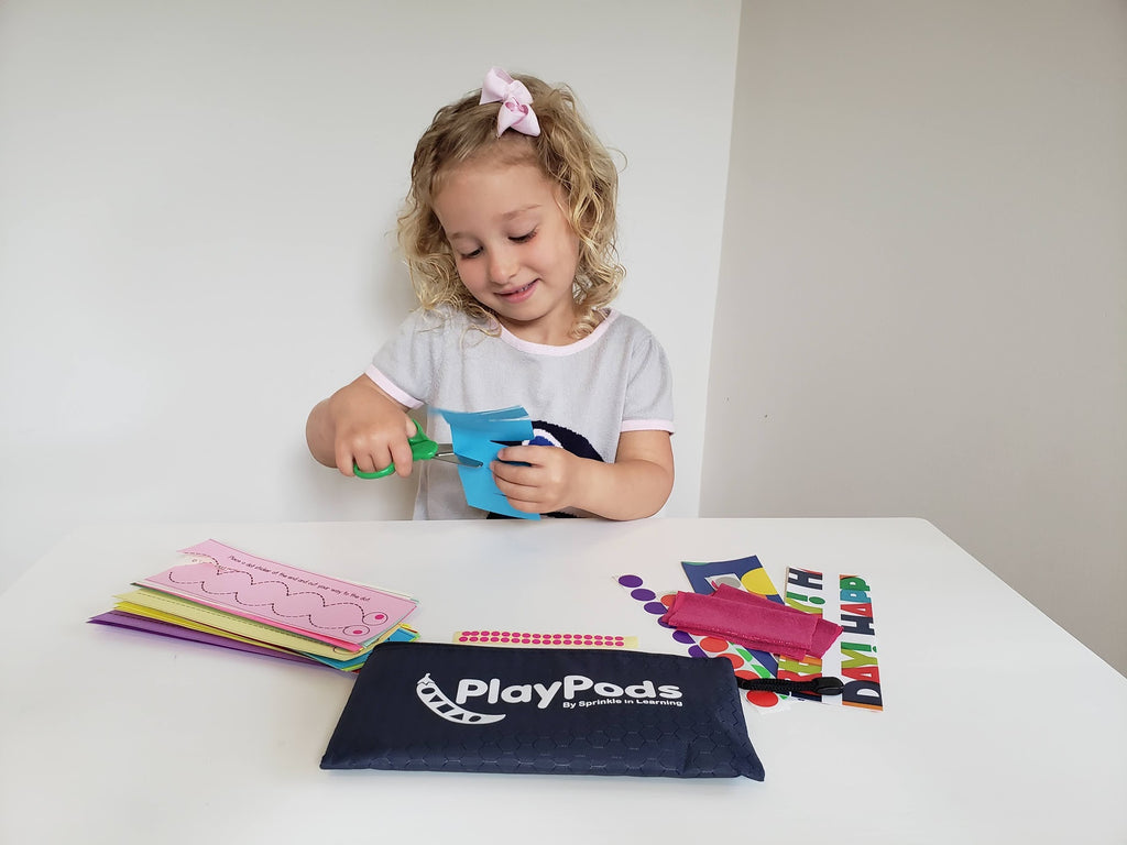 Little girl cutting blue paper with a green scissors and navy PlayPod contents on table