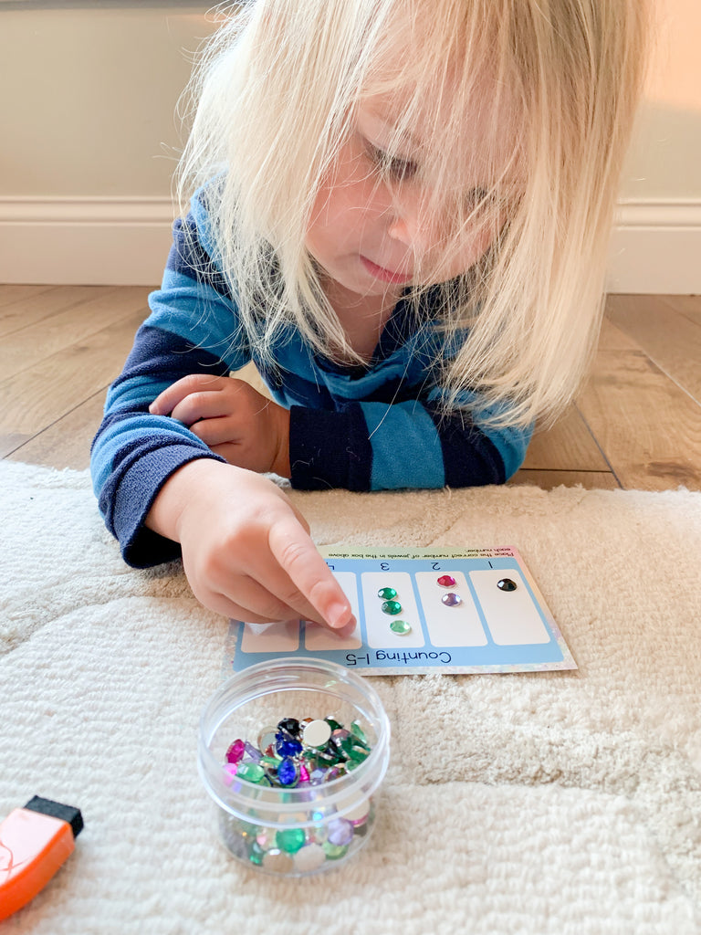 Little girl on floor using gems to count on instruction card